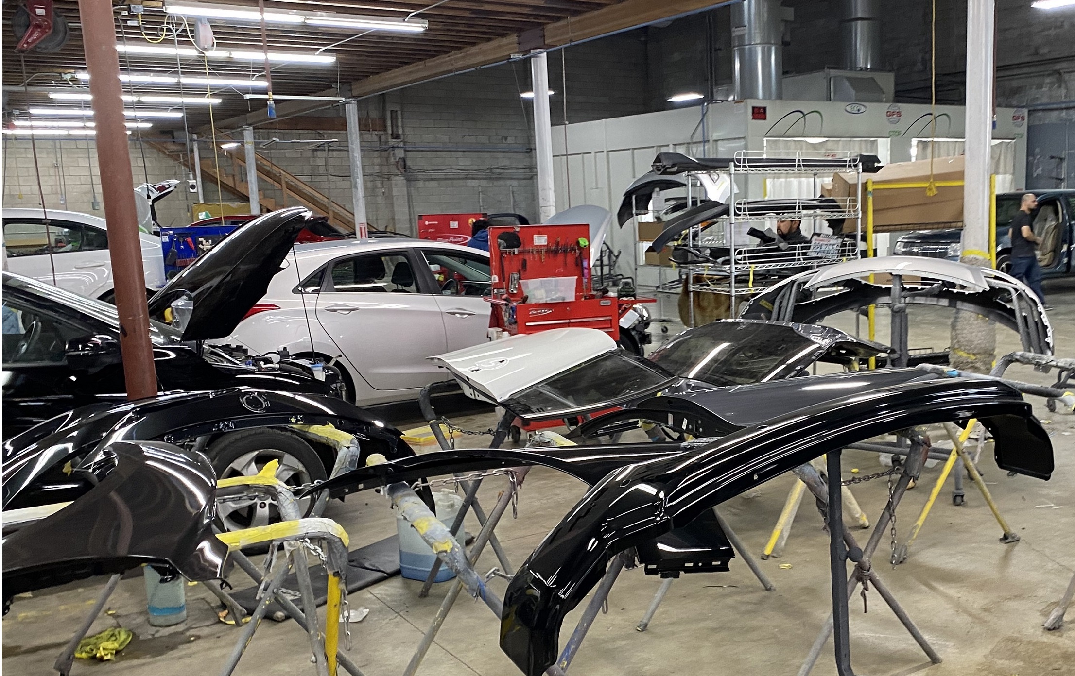 Different car parts seen in the foreground with vehicles in the background inside an autobody shop