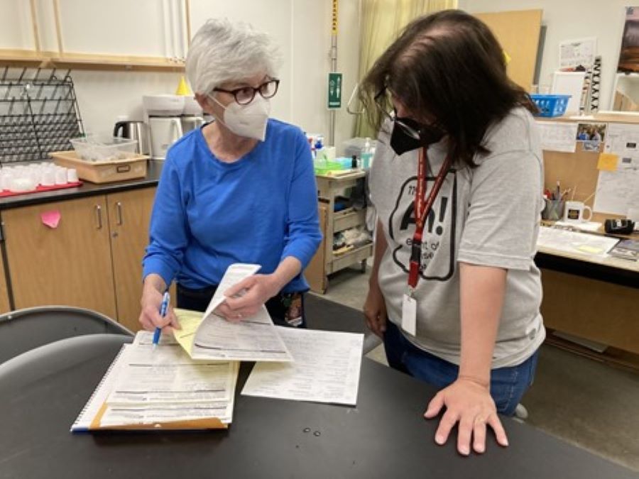A woman in a mask shows papers on a table to another person wearing a mask in a school classroom