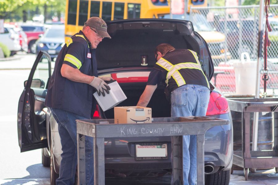 Two men wearing safety vests collect boxes of hazardous waste from the back trunk of a car