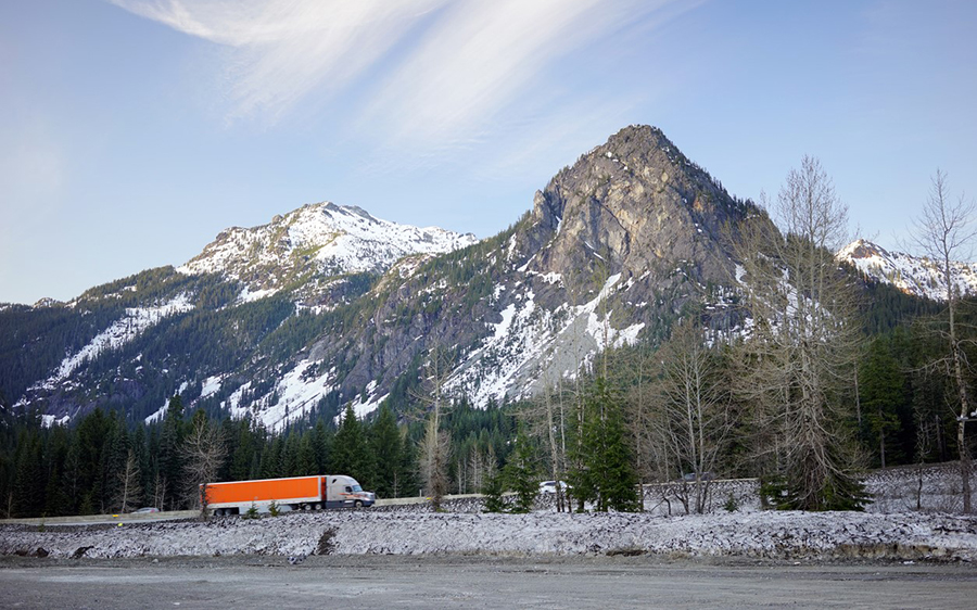 An orange semitruck on the road during winter time in front of a mountain range covered in snow